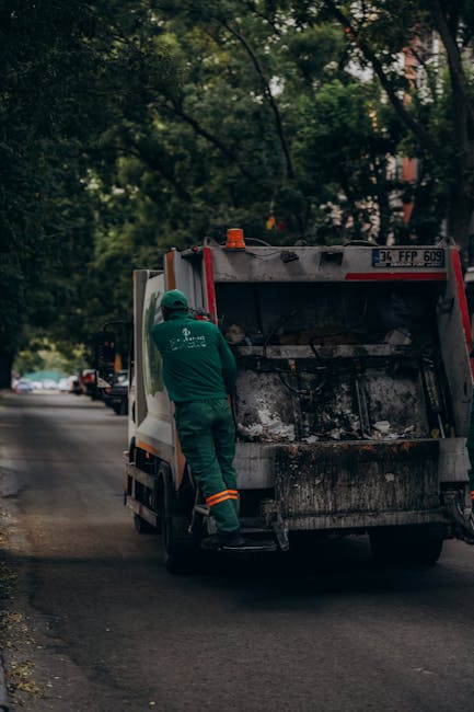 In a public outdoor space, a cluttered collection of overflowing rubbish bins and scattered waste surrounds a grey mixed paper and card recycling container labeled with a blue recycling symbol. Severa