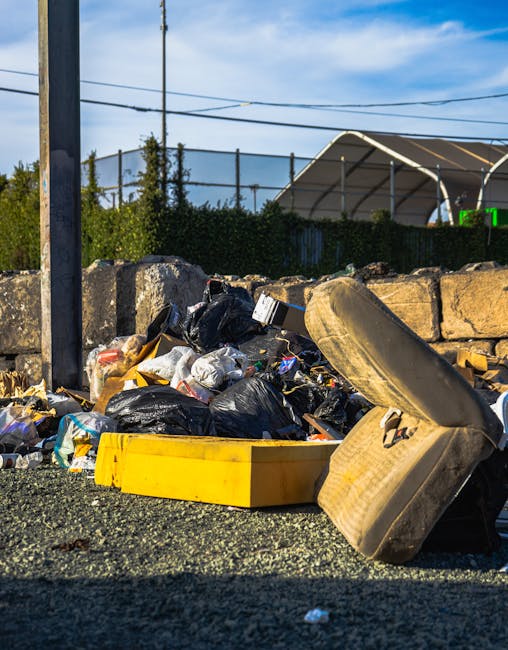 A large collection of mixed household waste and rubbish bags, including black and black-and-white plastic bags, cardboard boxes, and loose paper and packaging, are piled around a grey recycling bin la