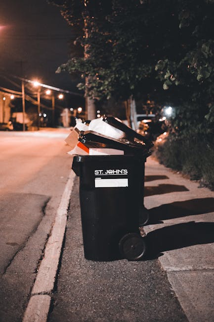 A row of large, rectangular wheelie bins positioned along a paved walkway beside a brick wall, with alternating green and blue colours. The green bins are closest in the foreground, featuring ridged,