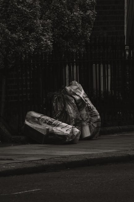 Two tightly packed black plastic garbage bags and a large cardboard box, all appearing to contain waste, are placed on the curbside pavement in front of a tall metal fence with vertical bars. The bags