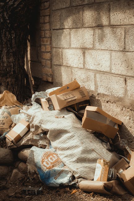 Two tightly packed black plastic garbage bags and a large cardboard box, all appearing to contain waste, are placed on the curbside pavement in front of a tall metal fence with vertical bars. The bags