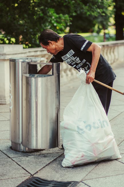 A man with dark hair is seen bending over a metal waste bin, picking up a piece of rubbish with his right hand while holding a large white plastic bag in his left hand. He is dressed in a black T-shir